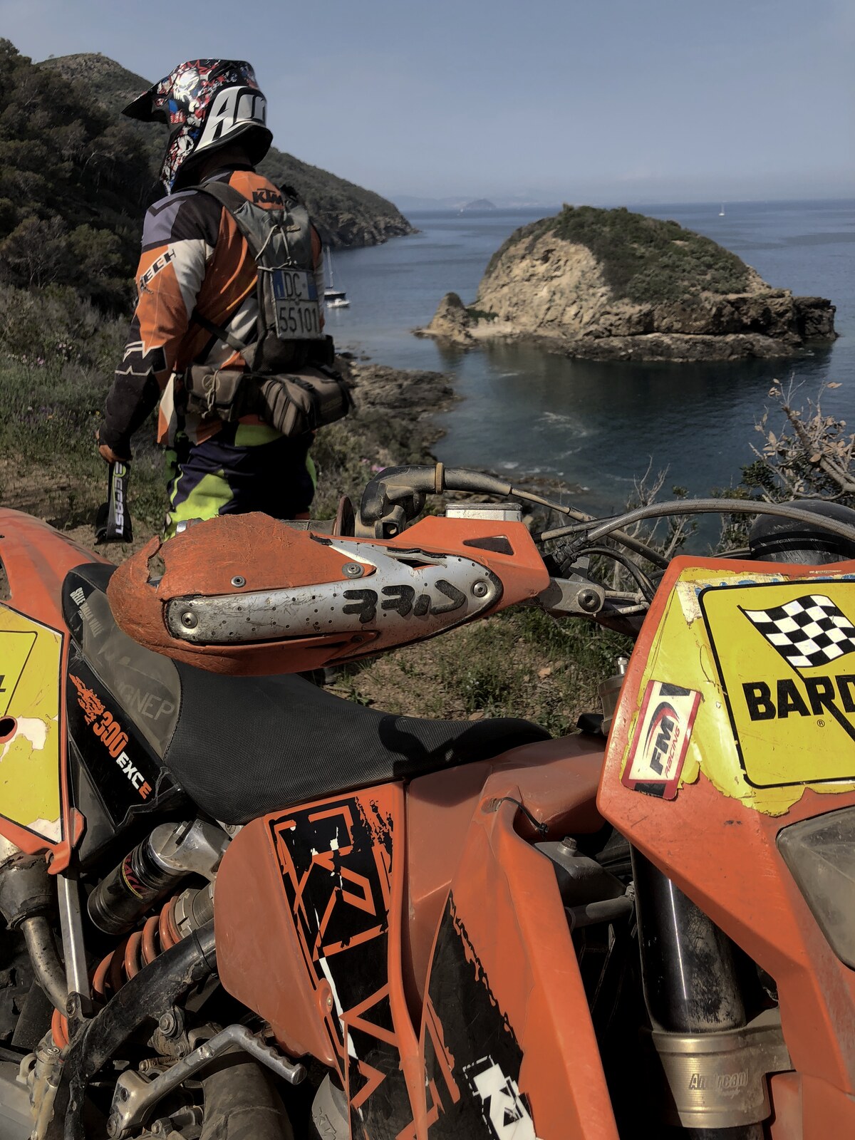 Enduro rider paused above a bay on Elba, scarred handguards in the foreground — all of us could ride